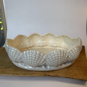 May include: An oval ceramic bowl with a scalloped rim and a textured exterior featuring a repeating shell pattern. The bowl has a glossy, off-white finish with subtle iridescent highlights that catch the light along the curved edges.
