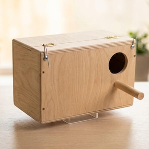 Peut inclure: Un nichoir rectangulaire en bois clair doté d'un trou d'entrée circulaire sur la façade, d'un perchoir cylindrique en bois qui dépasse et de deux charnières métalliques sur le couvercle supérieur fixées par des attaches en métal argenté.