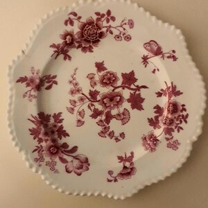May include: A white ceramic plate with a scalloped edge featuring a dark pink floral pattern. The design includes various clusters of flowers and leaves distributed across the surface, along with a small butterfly illustration on the right side.