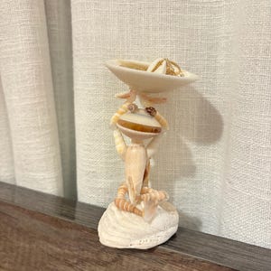 May include: A decorative figurine crafted from various seashells, featuring a creature-like form holding a small, shallow shell bowl above its head. The structure is mounted on a larger, flat white shell base, with gold-toned jewelry inside the bowl.
