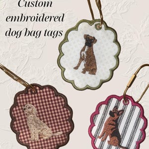 May include: Three scalloped-edge fabric bag tags with gold-tone metal clips. Each tag features an embroidered dog: a brown boxer on cream fabric, a light-colored doodle on red gingham, and a German shepherd on white and black striped fabric.