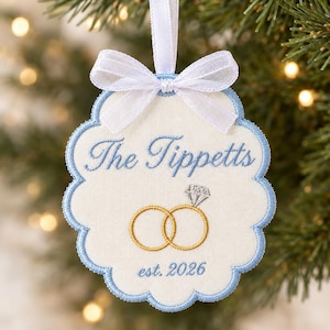 May include: A scalloped, off-white fabric ornament with light blue embroidery trim and a white ribbon bow. It features the embroidered text "The Tippetts" and "est. 2026" in blue, along with a graphic of two gold interlocking rings with a diamond.