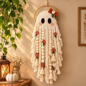 May include: A cream-coloured crocheted ghost wall hanging suspended from a wooden hoop. The ghost features black oval eyes, pink cheeks, and an orange bow with a white flower. It is decorated with small crocheted flowers and hanging tassels.