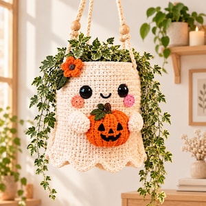 May include: A cream-coloured crochet ghost plant hanger featuring a smiling face with black eyes and pink cheeks. The ghost holds a small orange jack-o'-lantern with a green stem and has an orange flower accent. It hangs from a macrame cord.