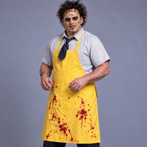 Peut inclure: Un tablier jaune avec des éclaboussures rouges ressemblant à du sang, porté sur une chemise à manches courtes rayée blanc et gris avec une cravate bleu foncé. L'ensemble est complété par un masque avec des détails cousus et du maquillage.