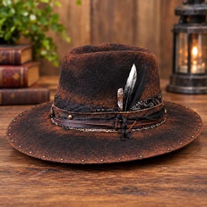Könnte beinhalten: Ein abgenutzter brauner Filzhut im Fedora-Stil mit einer breiten Krempe, die am Rand mit kleinen Metallnieten besetzt ist. Der Kopfbereich ist mit einem dunklen Stoffband, einer dünnen Lederschnur, einer Metallkette und drei Federn verziert.