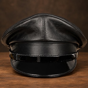 May include: A black leather cap featuring a structured crown with visible stitching and a wide, smooth leather band. The front includes a glossy, patent-finish visor and a thin, matching patent strap secured with small side buttons across the front.