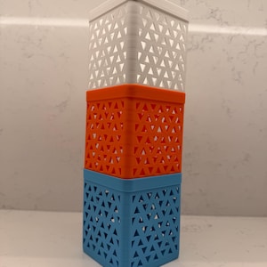 May include: A vertical stack of three square, hollow containers with a geometric triangular lattice pattern. The top container is white, the middle is orange, and the bottom is blue. Each section features a consistent open-work design.