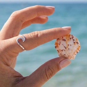 May include: A silver ring featuring a stylized wave design worn on a finger, positioned next to a small, fan-shaped seashell with a white base and brown speckled markings. The ring has a smooth, polished metallic finish and a curved, open-loop shape.