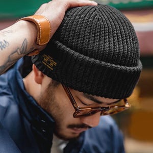 May include: A black ribbed knit beanie with a folded cuff. A small rectangular black label with gold text reading "VINTAGE CLOTHING" and "UNION WEAR CO" is stitched onto the side of the cuff. The beanie features a thick, textured vertical knit pattern.