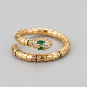 Puede incluir: Un anillo de serpiente en tono dorado con un patrón de escamas texturizado a lo largo de la banda. La cabeza de la serpiente está decorada con dos piedras verdes de corte marquesa y pequeños cristales transparentes. Piedras de colores en el cuerpo.