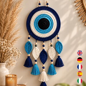 May include: A circular wall hanging featuring a crocheted evil eye design in concentric rings of black, light blue, white, and dark blue. The piece includes hanging strands decorated with wooden beads and crocheted leaf shapes in blue and white.