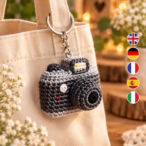 May include: A crochet camera keyring featuring a dark grey and light grey yarn body with a prominent circular lens, a small white button detail, and a silver metal ring attached to a beige canvas tote bag handle for secure hanging.