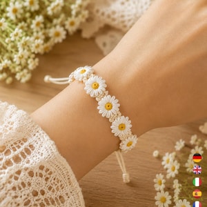 May include: A woven bracelet featuring a series of white daisy flowers with yellow centers, connected by a thin cream-colored cord. The bracelet has an adjustable sliding knot closure with two dangling cord ends, creating a floral pattern.