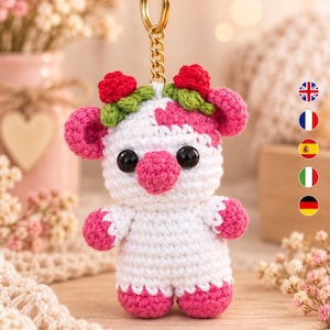 May include: A crochet cow keyring featuring a white body with pink patches, a pink snout, and black eyes. The cow is adorned with a crown of small red roses and green leaves on its head. It is attached to a gold-coloured metal ring and chain.