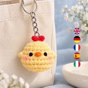 May include: A yellow crocheted chick keyring featuring a small red heart on top, two black bead eyes, an orange beak, and pink blush accents on the cheeks. The chick is attached to a silver-toned metal split ring and a short chain link connector.