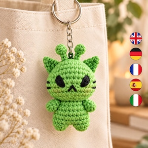 May include: A green crocheted keychain featuring a cat-like creature with two antennae, pointed ears, black almond-shaped eyes, and a small black stitched mouth. The figure is attached to a silver-toned metal keyring and chain.