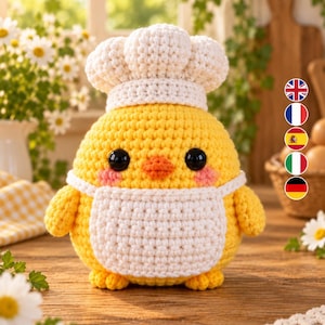 Può includere: Un pulcino all'uncinetto giallo che indossa un cappello da chef bianco e un grembiule bianco. Il pulcino ha grandi occhi neri, un piccolo becco arancione e guance rosa. La figura è rotonda e presenta una texture lavorata a maglia.
