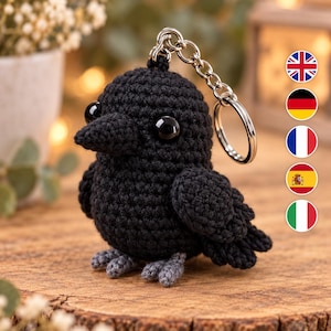 May include: A black crochet raven keychain featuring a round body, a pointed beak, and two large black plastic eyes. The bird has small grey feet and is attached to a silver-colored metal split ring by a short chain, creating a compact accessory.