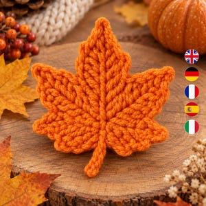 May include: A vibrant orange crochet maple leaf featuring a detailed central vein and textured stitch pattern. The leaf is crafted from yarn and displays a distinct stem at the base, showcasing a handmade construction with clear, defined loops.