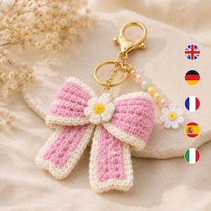 May include: A crochet keychain featuring a large pink bow with cream-colored edges, centered with a small white and yellow daisy flower. The gold-toned metal clasp is attached to a short strand of pastel-colored beads ending in a small white daisy.