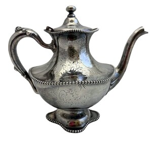 May include: A silver-plated teapot featuring an ornate, bulbous body with intricate floral engravings. It has a curved handle, a long spout, and a removable lid with a small finial. The base and rim are decorated with a beaded edge detail.