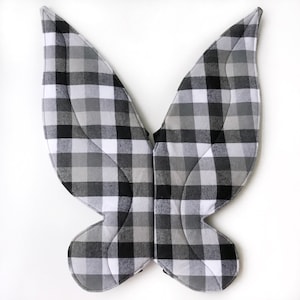 May include: Fairy wings in a black, white, and grey check pattern. The wings are made of quilted fabric and have a curved shape, with a central connecting piece. Ideal for costume or dress-up play, these wings are a fun accessory.