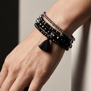 Puede incluir: Un conjunto de cuatro pulseras con cuentas negras facetadas, cuentas negras redondas y lisas, y una hilera de pequeñas cuentas metálicas plateadas. El set incluye un pequeño colgante de borla de tela negra en una de las pulseras.