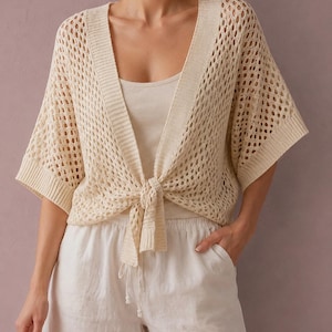 May include: A cream-colored, open-front knit cardigan featuring a loose, net-like crochet pattern, wide short sleeves, and a ribbed trim along the neckline and front edges that ties into a knot at the waist, worn over a simple cream tank top.