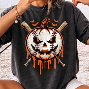 May include: A charcoal grey t-shirt featuring a graphic of a white baseball designed as a jack-o'-lantern with red stitching, sharp eyes, and a jagged mouth. Two crossed wooden baseball bats are behind the pumpkin, with orange liquid dripping down.
