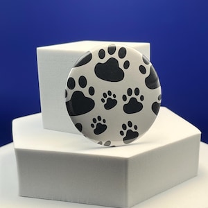 May include: A circular pin-back button featuring a repeating pattern of black paw prints on a light gray background. The design covers the entire surface of the round button, showcasing multiple sizes of animal paw prints arranged in a scattered layout.