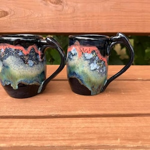 May include: Two ceramic mugs featuring a glossy black base that transitions into a layered glaze of vibrant coral, speckled blue, and sage green. Each mug has a curved handle and a smooth, rounded body with a reactive drip pattern.