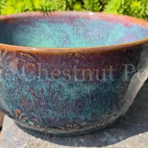 May include: A ceramic bowl featuring a reactive glaze with blended shades of deep teal, dark blue, and earthy brown. The exterior surface displays a subtle embossed floral pattern. The text Lynne Chestnut Pottery is visible across the center.