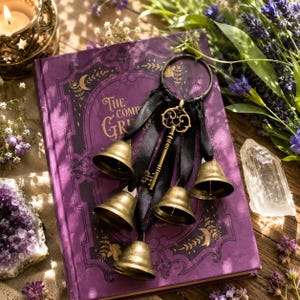 May include: A metal ring adorned with four brass-coloured bells and a decorative skeleton key, all suspended by black ribbons. The object rests on a purple hardcover book titled The Complete Grimm, featuring gold-coloured filigree and moon motifs.