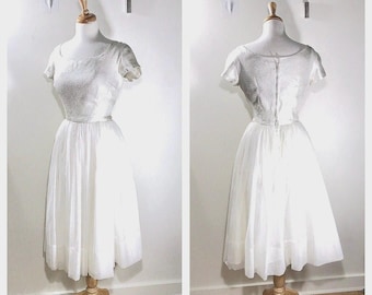 May include: A white dress with short sleeves and a textured bodice. The garment features a scoop neckline and a full, knee-length skirt made of layered fabric. The back of the dress displays a central vertical zipper closure extending from the neckline.