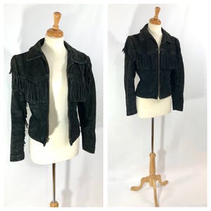 May include: A black suede jacket with fringe detailing on the shoulders and sleeves. The jacket has a zipper closure and a fitted silhouette.