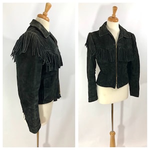 May include: A black suede jacket with fringe detailing on the shoulders, collar, and hem. The jacket has a zipper closure and a fitted silhouette.