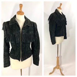May include: A black suede jacket with fringe detailing on the shoulders, hem, and sleeves. The jacket has a zipper closure and a collared neckline.