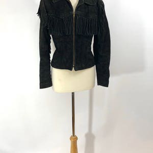 May include: A black suede jacket with fringe detailing on the shoulders and sleeves. The jacket has a zipper closure and a fitted silhouette.