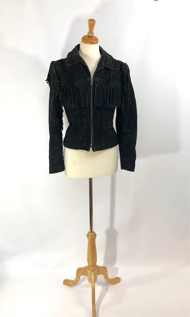 May include: A black suede jacket with fringe detailing on the shoulders and sleeves. The jacket has a zipper closure and a fitted silhouette.
