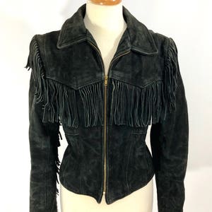 May include: A black suede jacket with fringe detailing on the shoulders, collar, and hem. The jacket has a zipper closure and a classic collar.