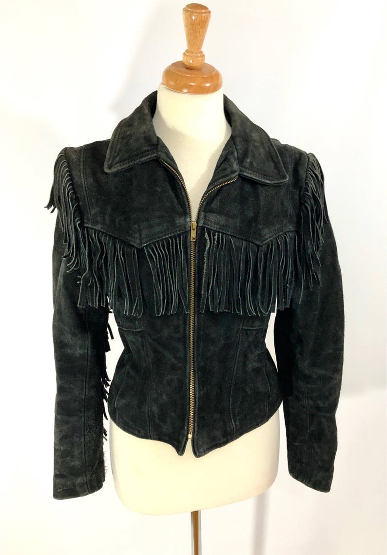 May include: A black suede jacket with fringe detailing on the shoulders, collar, and hem. The jacket has a zipper closure and a classic collar.