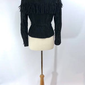 May include: Black suede jacket with fringe detailing on the sleeves and hem. The jacket is fitted and has a stand-up collar.