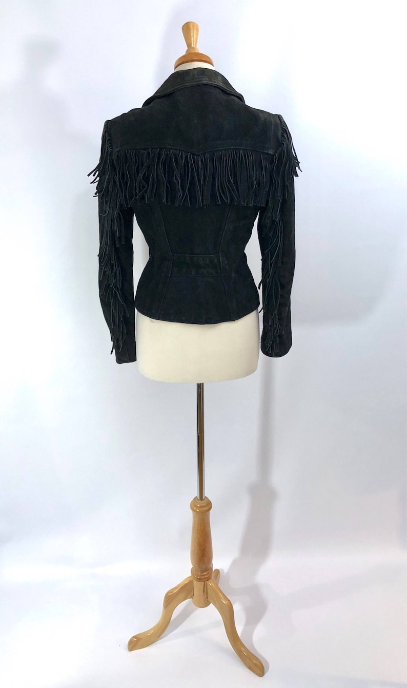 May include: Black suede jacket with fringe detailing on the sleeves and hem. The jacket is fitted and has a stand-up collar.