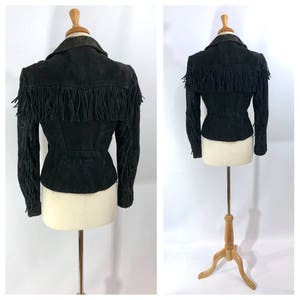 May include: Black suede jacket with fringe detailing on the sleeves and around the collar. The jacket has a fitted silhouette and a cinched waist.