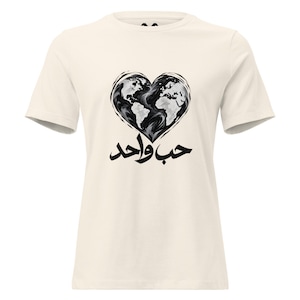 May include: A cream-colored short-sleeve t-shirt featuring a black and white graphic of a heart-shaped world map. Below the heart, the Arabic text "حب واحد" is printed in bold black calligraphy, centered on the chest of the garment.