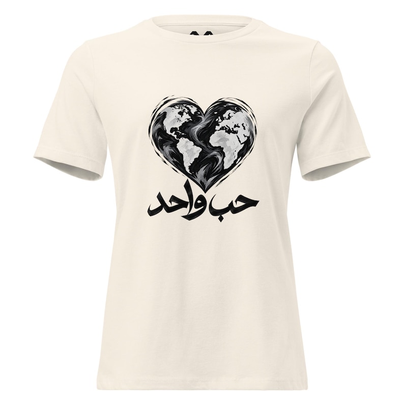 May include: A cream-colored short-sleeve t-shirt featuring a black and white graphic of a heart-shaped world map. Below the heart, the Arabic text "حب واحد" is printed in bold black calligraphy, centered on the chest of the garment.