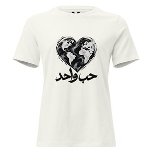 May include: An off-white short-sleeve t-shirt featuring a black and white graphic of a heart-shaped world map. Below the heart, the Arabic text "حب واحد" is printed in bold black calligraphy, centered on the chest of the garment.