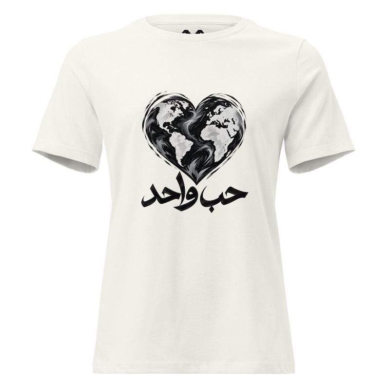 May include: An off-white short-sleeve t-shirt featuring a black and white graphic of a heart-shaped world map. Below the heart, the Arabic text "حب واحد" is printed in bold black calligraphy, centered on the chest of the garment.