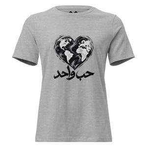 May include: A heather gray short-sleeve t-shirt featuring a black and white graphic of a heart-shaped world map. Below the heart, the Arabic text "حب واحد" is printed in a bold, black, stylized calligraphic font across the chest area.